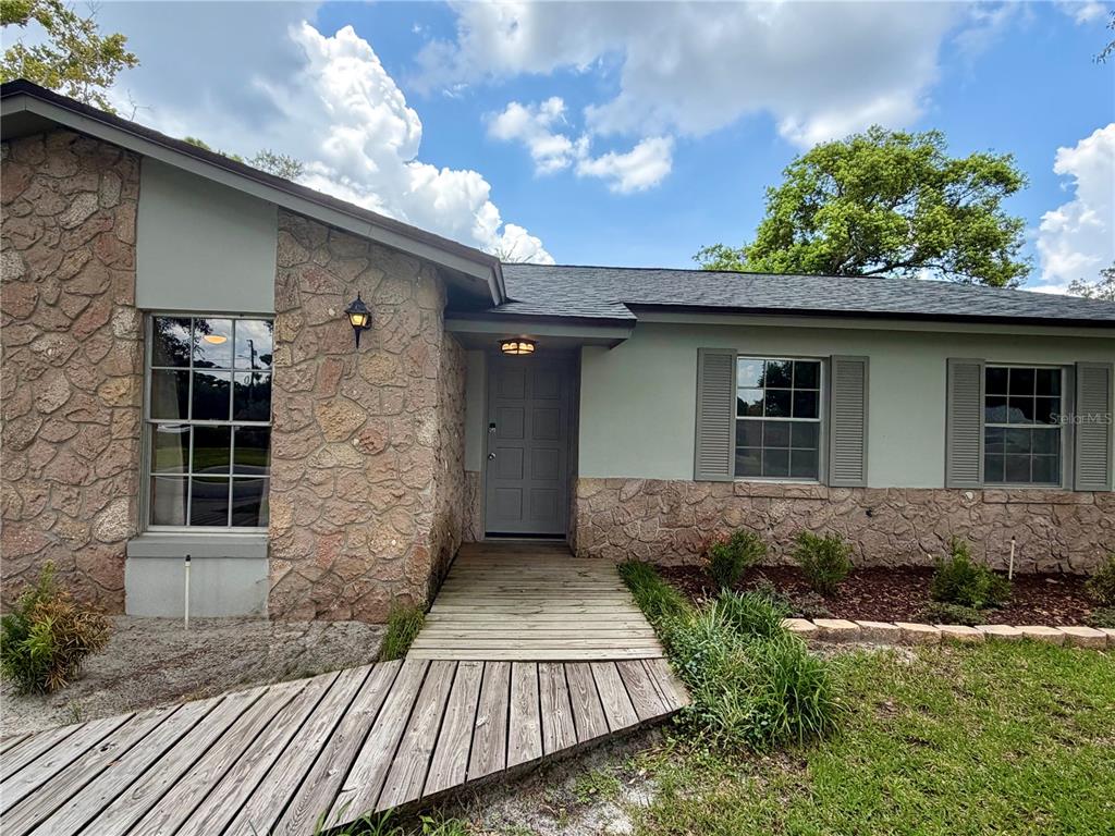 A single-story house exterior features stone siding, a wooden walkway, and a gray front door — 1 KING ARTHUR COURT, CASSELBERRY, FL