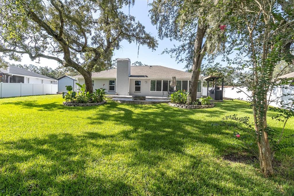 A backyard view of a single-story house with a large lawn and two mature trees — 8345 APPLE ORCHARD ROAD, SPRING HILL, FL