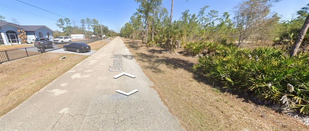 A paved road stretches past a residential house and a wooded area with palmettos — 1002 GLENN AVENUE, LEHIGH ACRES, FL