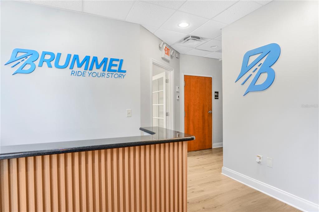 An office reception area features a wooden desk, light wood flooring, and blue company signage — 8615 COMMODITY CIRCLE UNIT 2, ORLANDO, FL