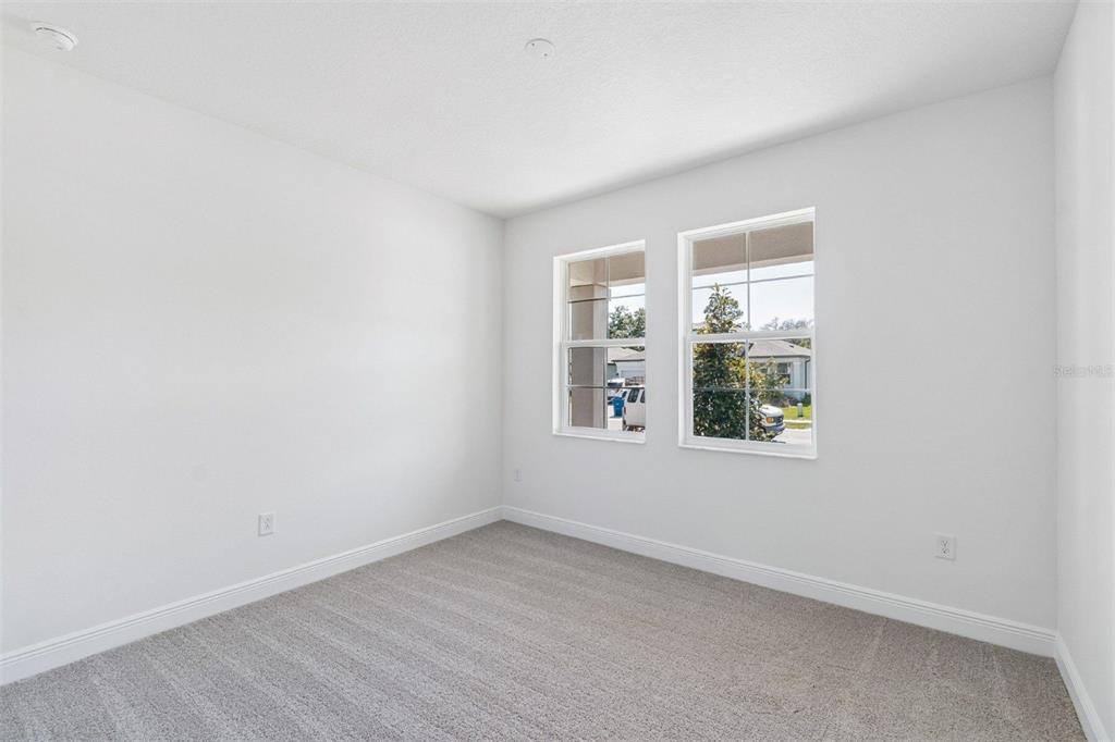 An empty bedroom features white walls, gray carpeting, and two windows overlooking a suburban street — 2210 CLARY SAGE DRIVE, SPRING HILL, FL