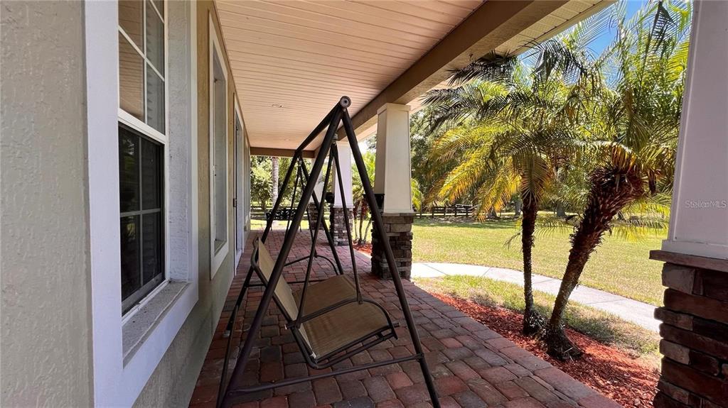 A covered porch features a metal swing, brick flooring, and views of a grassy yard — 3060 RAMBLER AVENUE, ST CLOUD, FL
