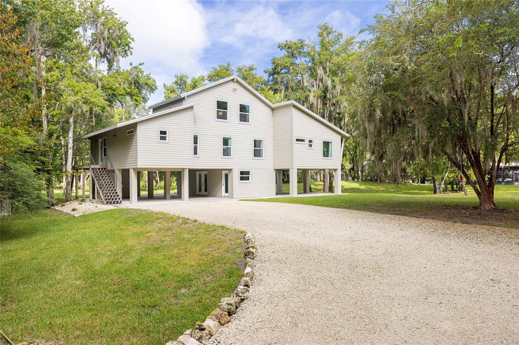 A light-colored, elevated two-story house with a gravel driveway and surrounding trees — 11772 N RIVER SOUND PATH, INGLIS, FL