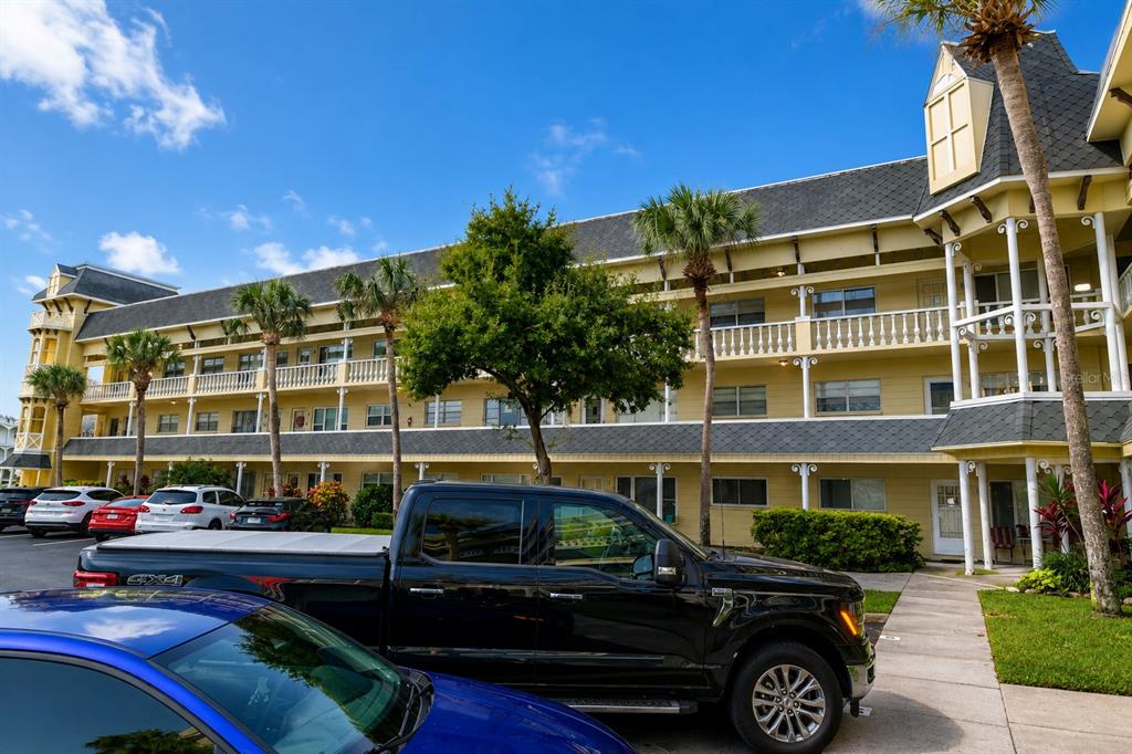 A multi-story yellow apartment building with balconies and palm trees sits behind parked cars — 2459 FRANCISCAN DRIVE UNIT 37, CLEARWATER, FL