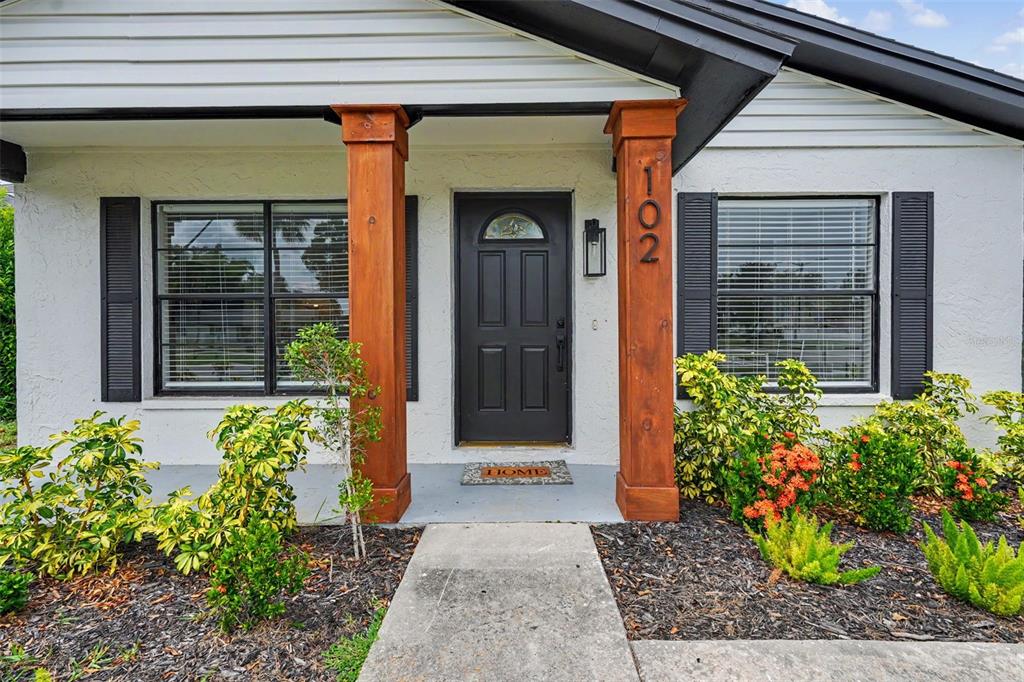 The front exterior of a house features a dark door, wooden pillars, and landscaped garden beds — 102 SHORE DRIVE E, OLDSMAR, FL