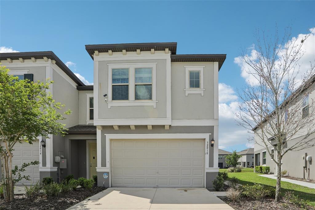 A two-story townhouse features a beige stucco exterior, a two-car garage, and a small lawn — 32010 BLUE PASSING LOOP, WESLEY CHAPEL, FL