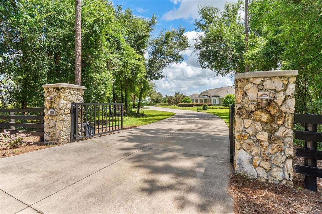 A concrete driveway leads through a stone-pillared gate toward a house surrounded by trees — 10615 NE 104TH CIRCLE, OXFORD, FL