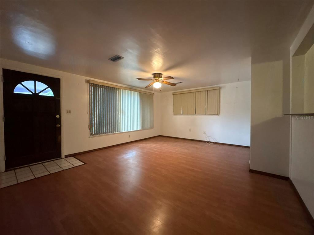 An empty living room features wood-look flooring, a ceiling fan, and vertical window blinds — 671 DEXTER DRIVE, DUNEDIN, FL