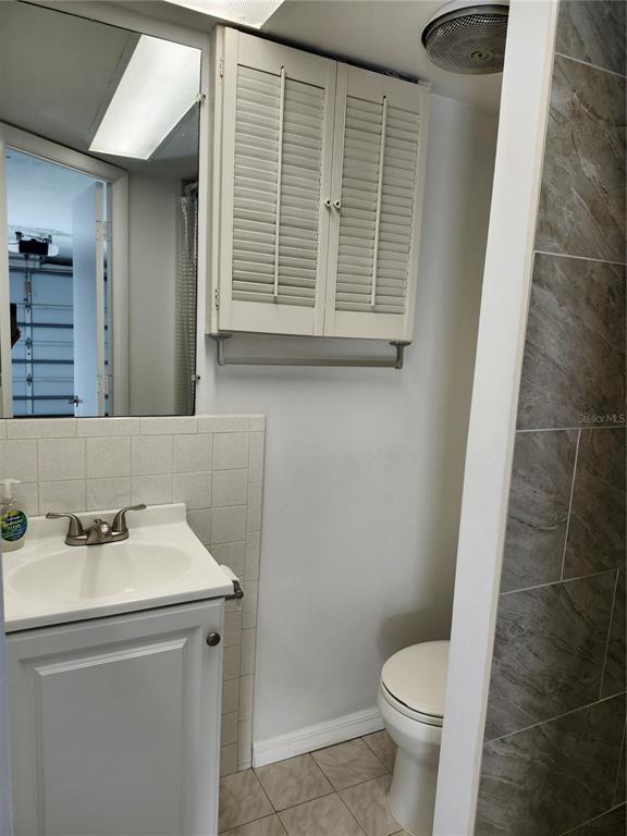 A bathroom features a white vanity, a wall-mounted cabinet, a toilet, and gray tiled walls — 6144 SAPPHIRE DRIVE, NEW PORT RICHEY, FL
