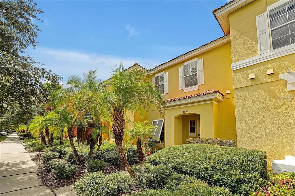 A yellow two-story townhouse exterior features palm trees, manicured hedges, and a paved sidewalk — 8591 BAY LILLY LOOP, KISSIMMEE, FL