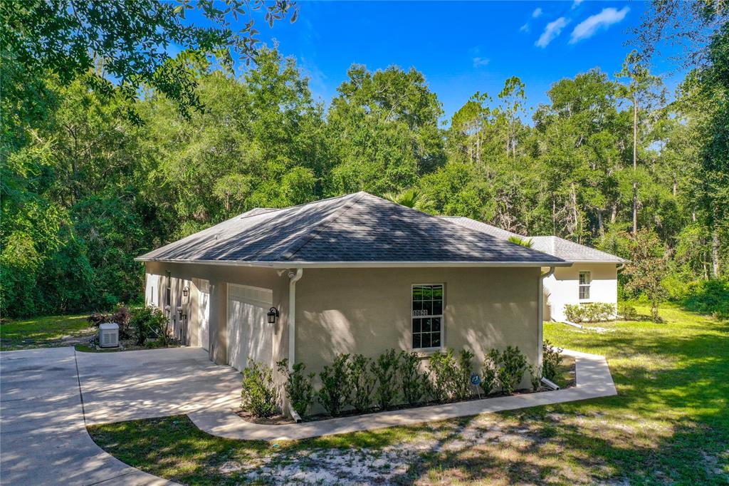 An exterior view of a tan house with a garage and a surrounding wooded area — 10620 NE 104TH CIRCLE, OXFORD, FL