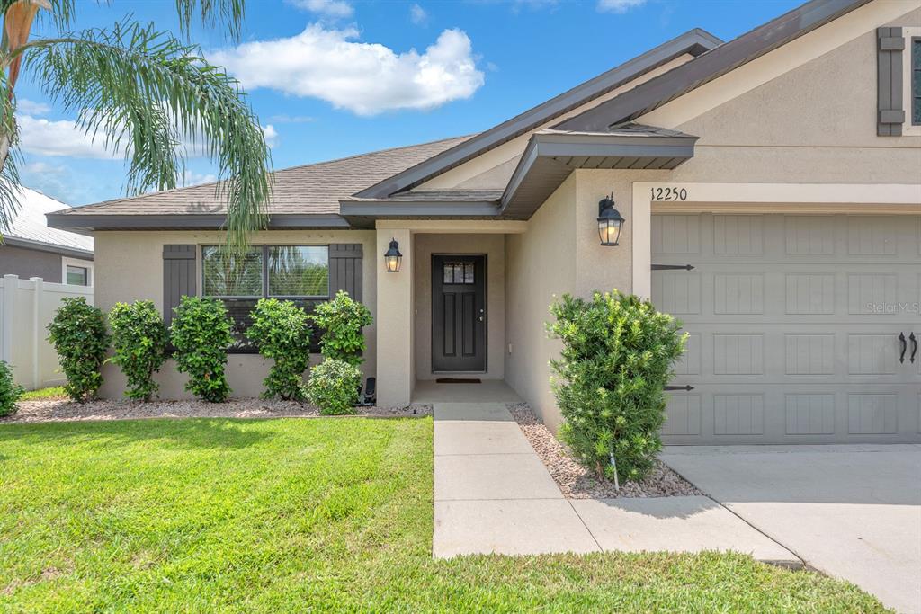 A single-story house exterior features a gray garage, a dark front door, and manicured landscaping — 12250 LEGACY BRIGHT STREET, RIVERVIEW, FL
