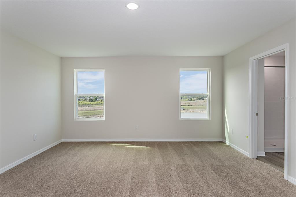 An empty bedroom features beige carpet, light-colored walls, two windows, and an adjacent bathroom doorway — 36715 THORNHEDGE DRIVE, ZEPHYRHILLS, FL