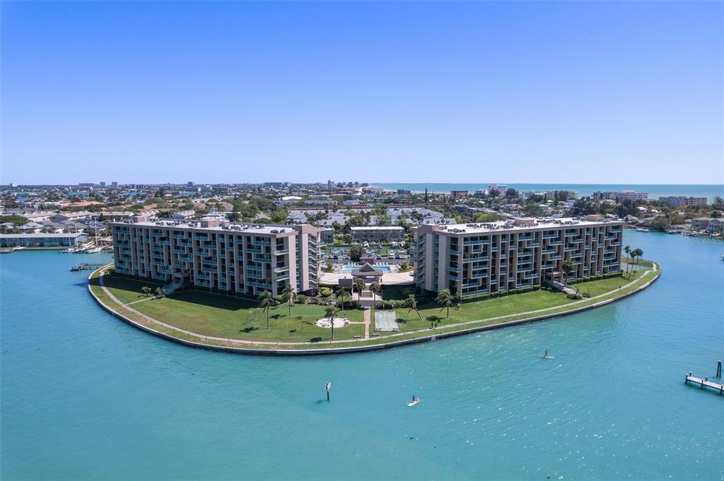 An aerial view of a multi-story apartment complex situated on a grassy peninsula surrounded by water — 1 KEY CAPRI UNIT 303E, TREASURE ISLAND, FL
