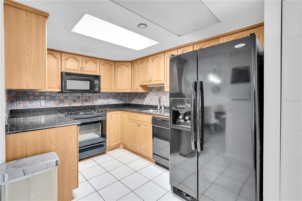 A kitchen features light wood cabinets, black appliances, dark countertops, and white tile flooring — 1 KEY CAPRI UNIT 303E, TREASURE ISLAND, FL