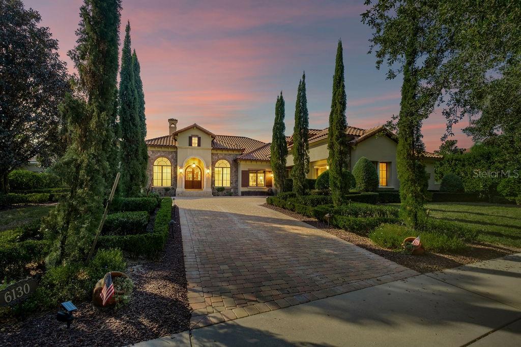 A Mediterranean-style house with a brick driveway and tall cypress trees at sunset — 6130 GROSVENOR SHORE DRIVE, WINDERMERE, FL