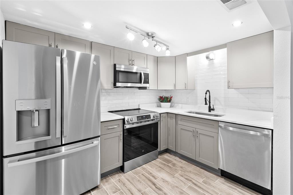 A modern kitchen features stainless steel appliances, grey cabinetry, white countertops, and wood-look tile flooring — 505 PLAZA SEVILLE COURT UNIT 9, TREASURE ISLAND, FL