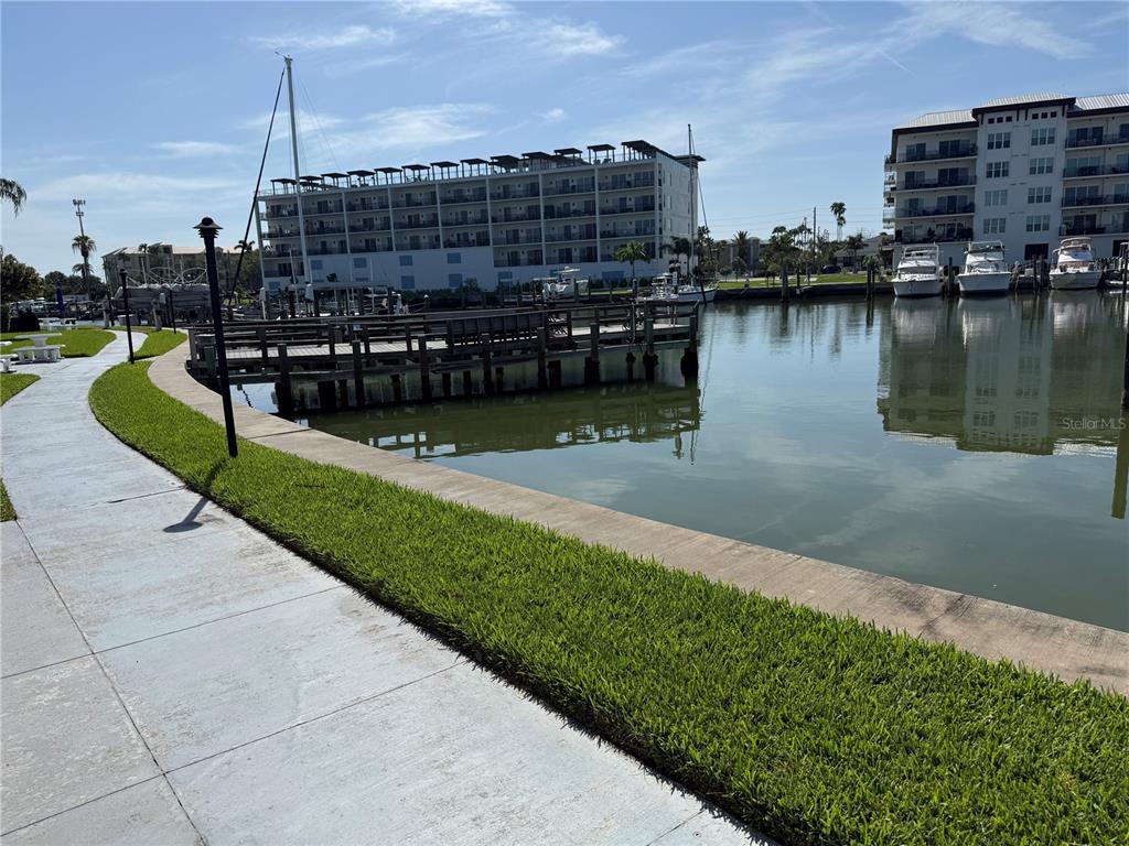 A paved walkway borders a grassy lawn, a wooden dock, and a canal with boats — 179 MEDALLION BOULEVARD UNIT D, MADEIRA BEACH, FL