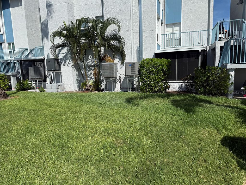 The exterior of a multi-story residential building features a grassy lawn, palm trees, and HVAC units — 179 MEDALLION BOULEVARD UNIT D, MADEIRA BEACH, FL