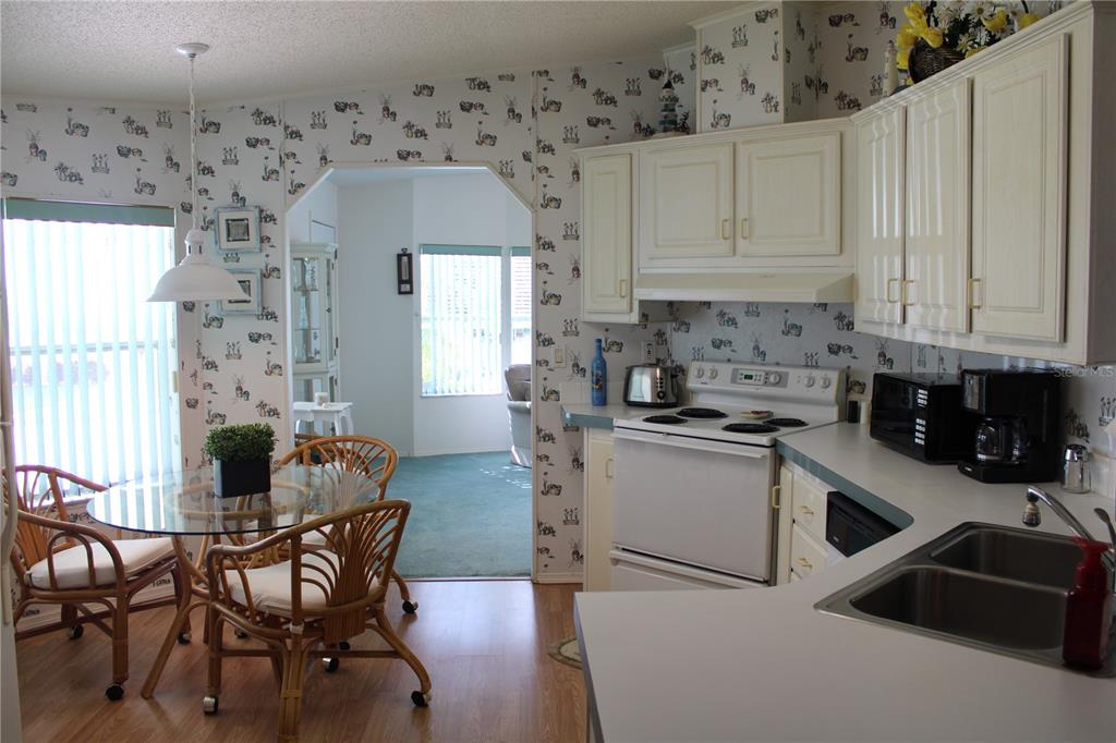 A kitchen with white cabinets and patterned wallpaper opens to a dining area with wicker chairs — 8314 NANCY LANE, ELLENTON, FL