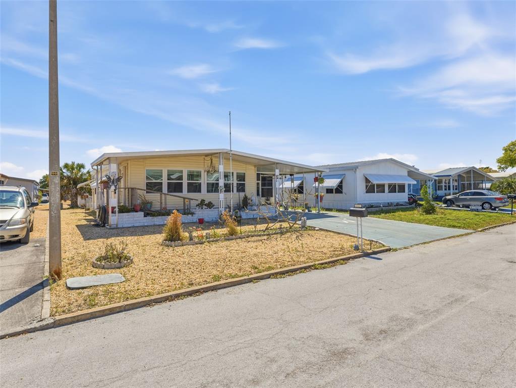 A light yellow manufactured home with a carport and gravel yard under a blue sky — 7402 CASCADE DRIVE, HUDSON, FL