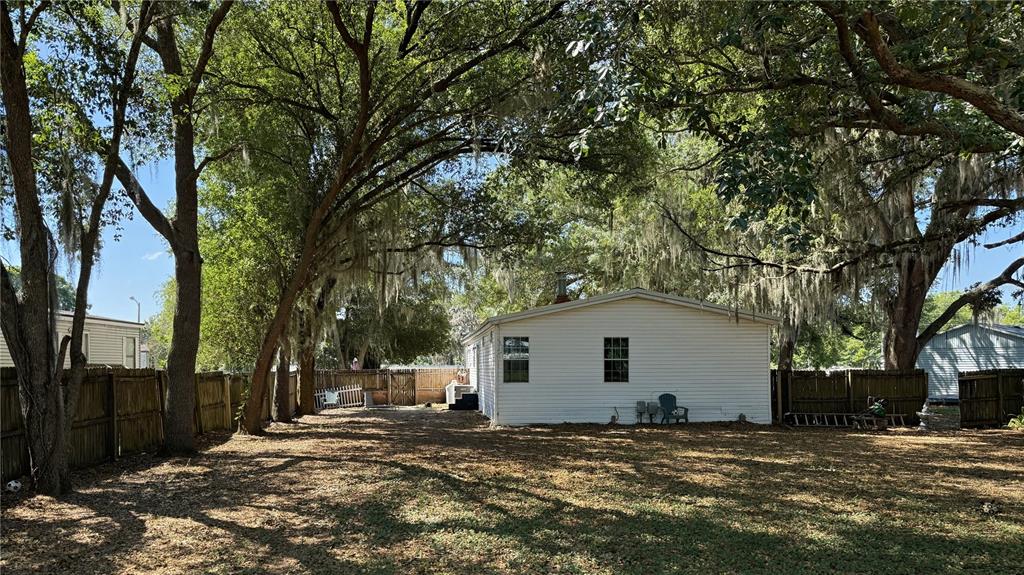 A white house sits in a large, shaded backyard with mature trees and a wooden fence — 4817 ROLLINGLEN LOOP W, LAKELAND, FL