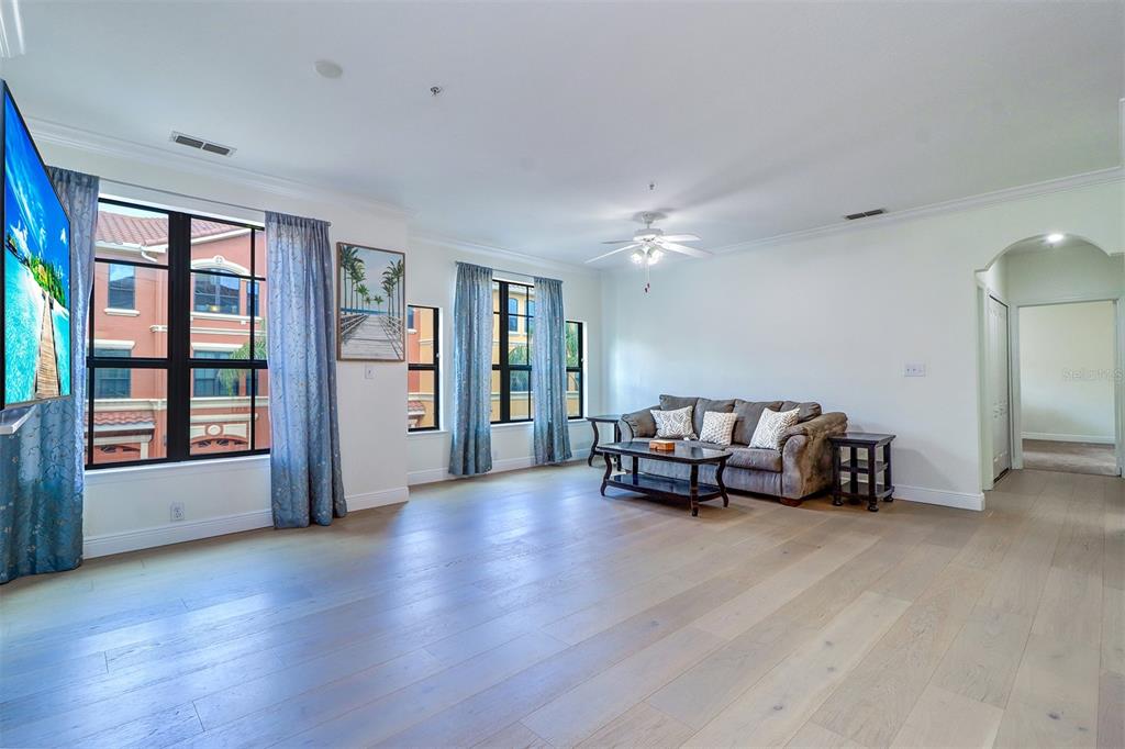 A living room with light wood floors, a brown sofa, and large windows with blue curtains — 2741 VIA CIPRIANI UNIT 922B, CLEARWATER, FL