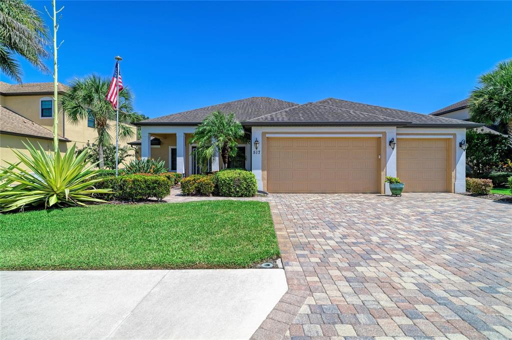 A single-story house with a tan garage, paver driveway, and American flag on a sunny day — 517 CHANTILLY TRAIL, BRADENTON, FL