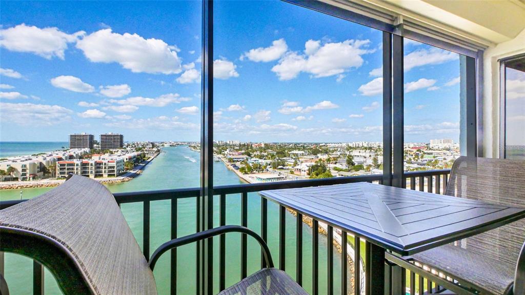 A balcony with outdoor furniture overlooks a waterway, buildings, and the ocean under a sky — 7150 SUNSET UNIT 903E, ST PETE BEACH, FL