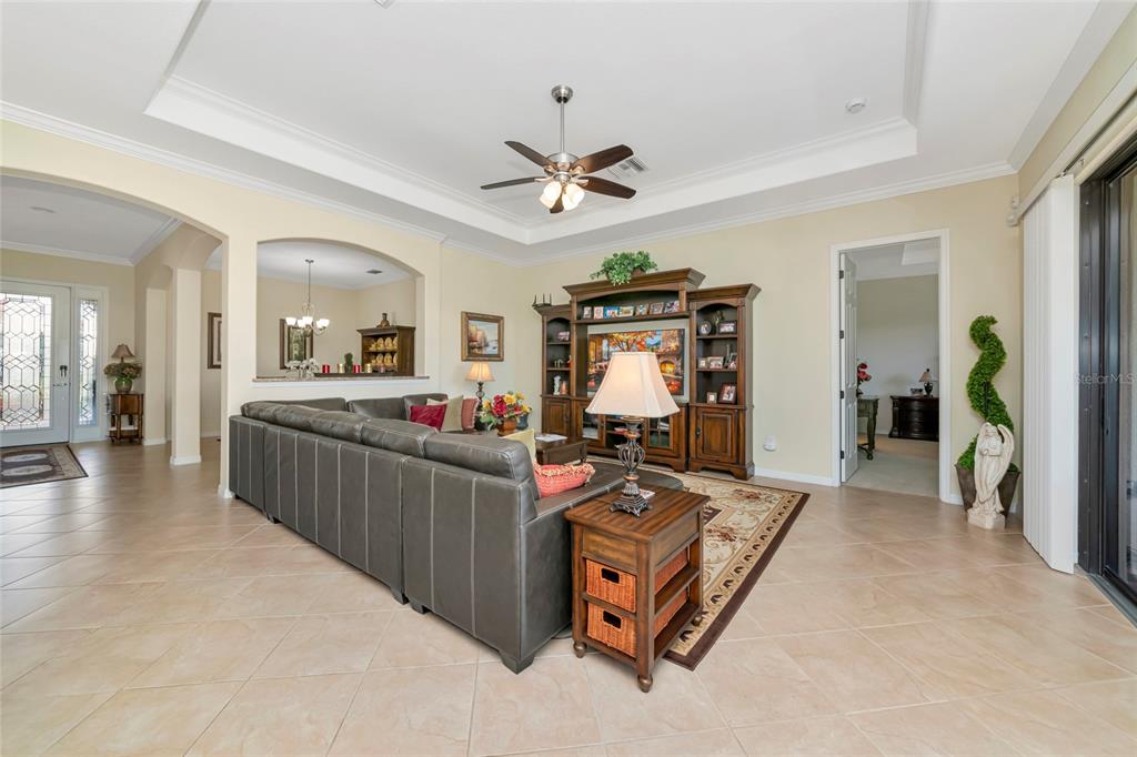 A living room features a tray ceiling, tile flooring, a large sectional, and an entertainment center — 13026 RAMBLEWOOD TRAIL, BRADENTON, FL