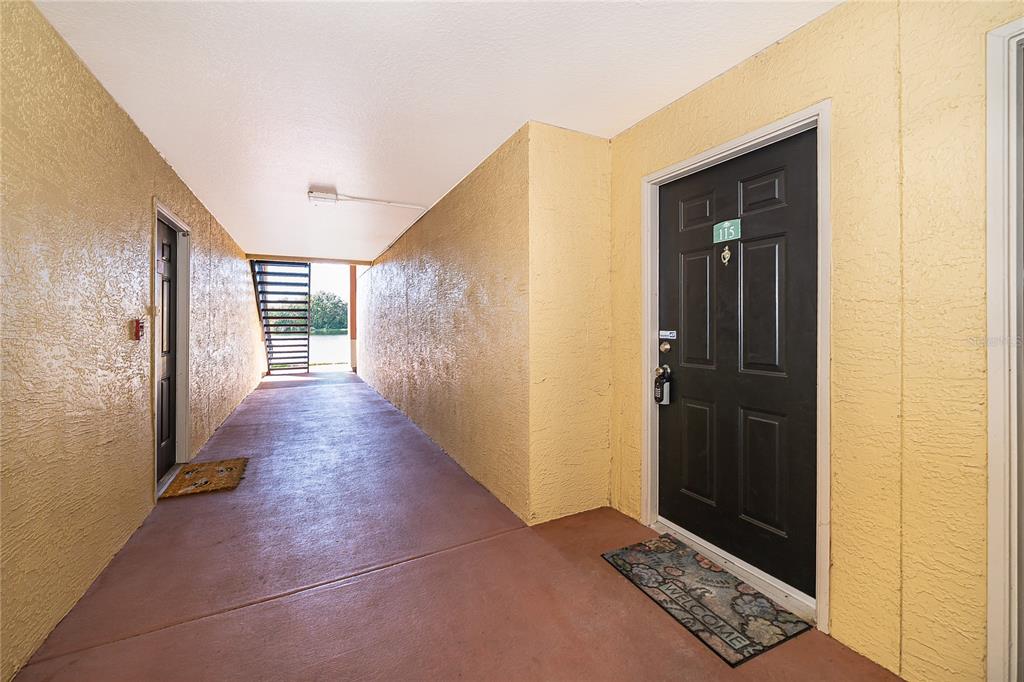 An outdoor apartment hallway features yellow textured walls, a red concrete floor, and black doors — 1939 SUMMER CLUB DRIVE UNIT 115, OVIEDO, FL
