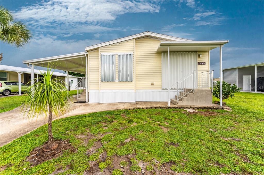 A yellow mobile home features a metal carport, concrete steps, and storm shutters on windows — 7464 DANVERS CIRCLE, PORT CHARLOTTE, FL