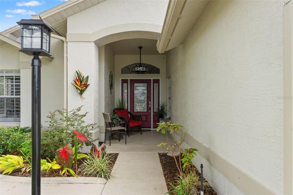 A covered entryway features a red front door, a chair, and a decorative wall hanging — 7350 ROSEMONT LANE, SPRING HILL, FL