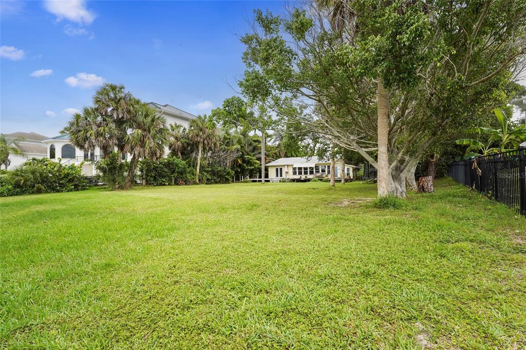 A large, grassy backyard features mature trees, tropical landscaping, and a distant single-story house — 7538 WESTMORELAND DRIVE, SARASOTA, FL