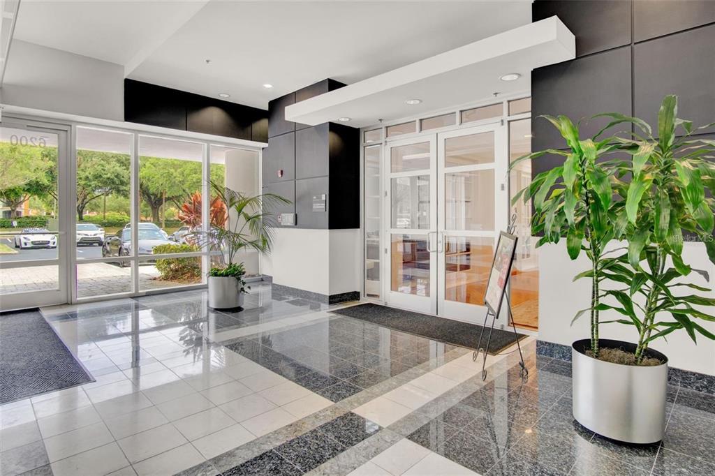 A modern building lobby features polished tile floors, glass entry doors, and two large potted plants — 6200 METROWEST BOULEVARD UNIT 103B, ORLANDO, FL