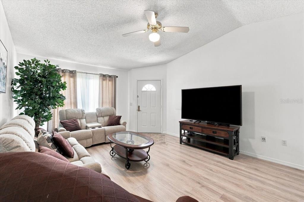 A living room features light wood flooring, a ceiling fan, a television, and neutral furniture — 1837 PRAIRIE DUNES CIRCLE S, LAKELAND, FL
