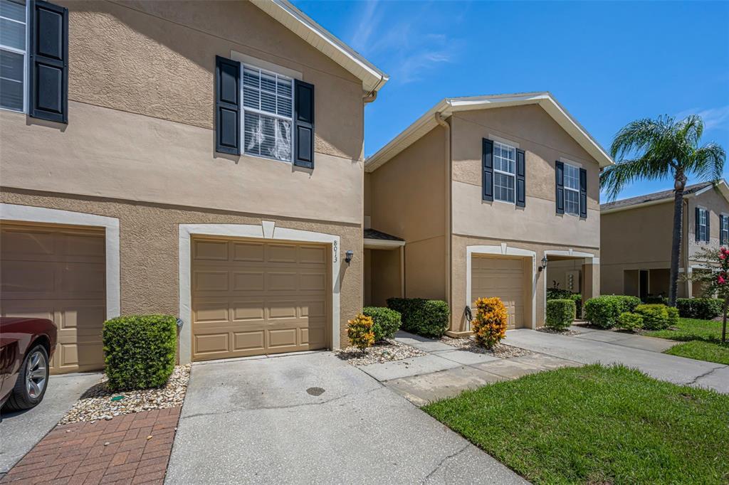 Two tan townhomes with attached garages and dark shutters sit under a clear blue sky — 8013 SUTTON TERRACE LANE, TAMPA, FL