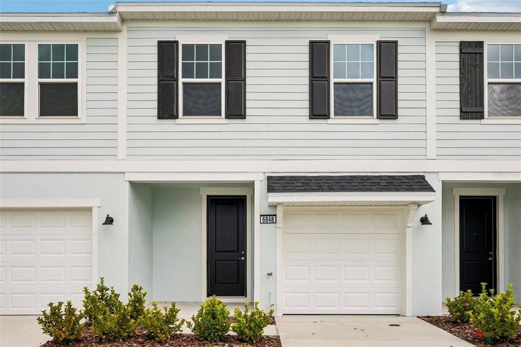 A two-story townhouse exterior features light blue siding, black shutters, and a single-car garage — 6848 HOLCOMBE DOWN DRIVE, ZEPHYRHILLS, FL