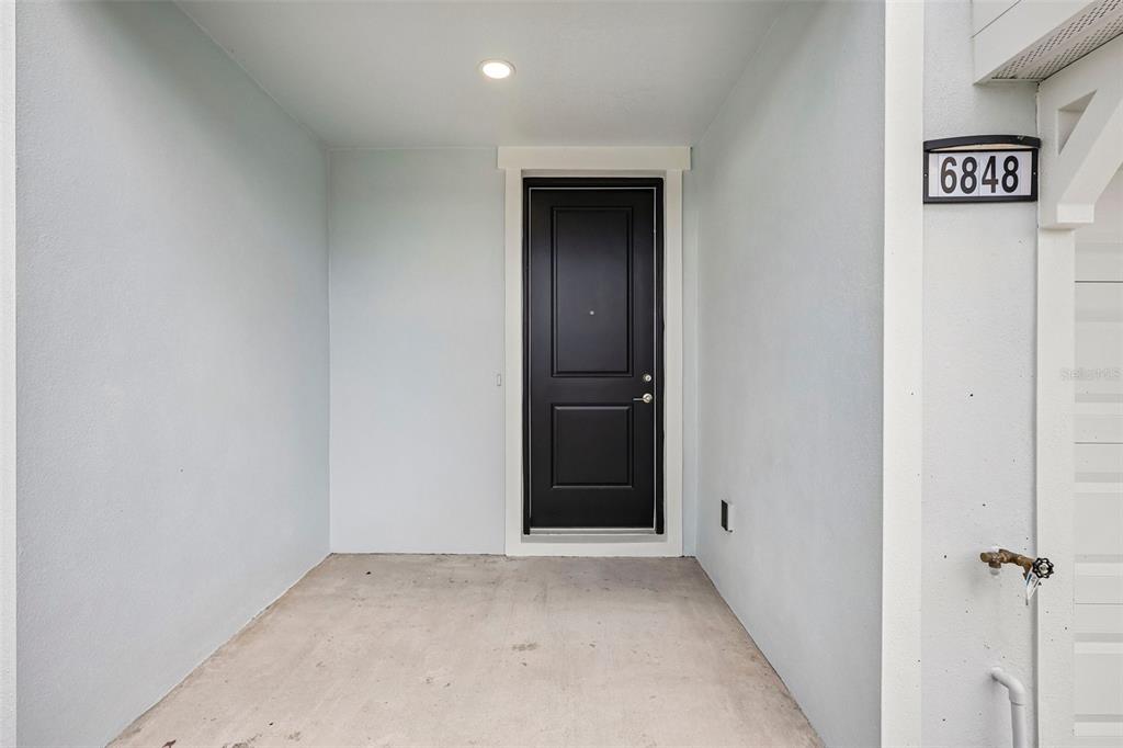 A black front door is set within a covered concrete entryway of a light-colored building — 6848 HOLCOMBE DOWN DRIVE, ZEPHYRHILLS, FL