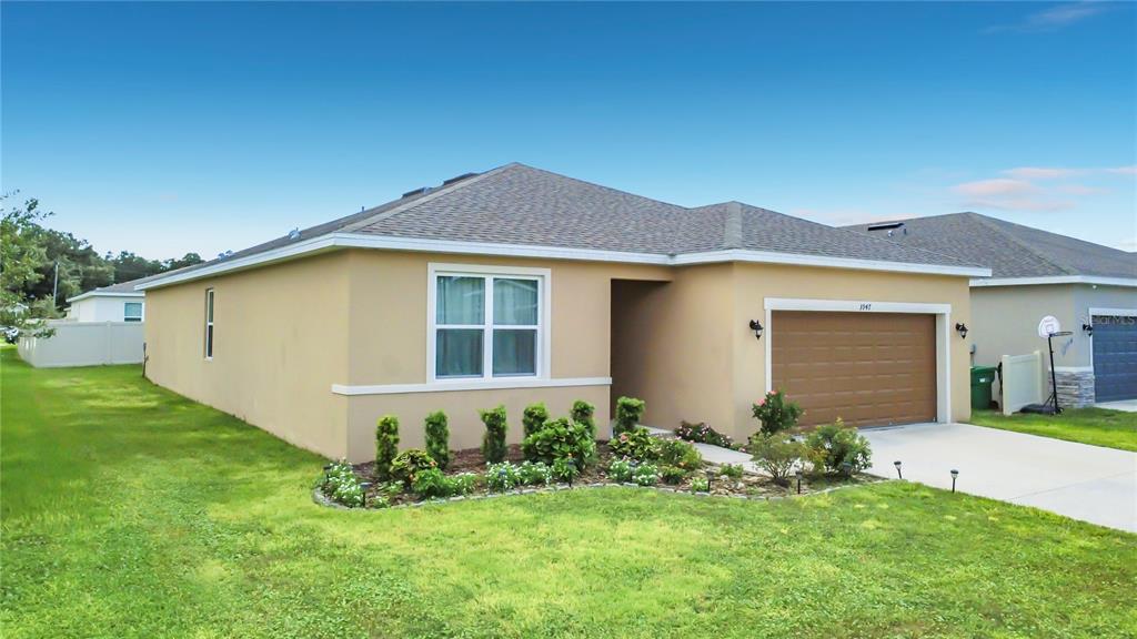 A single-story tan stucco house with a brown garage door and a landscaped front yard — 3947 MAYHILL LOOP, EUSTIS, FL