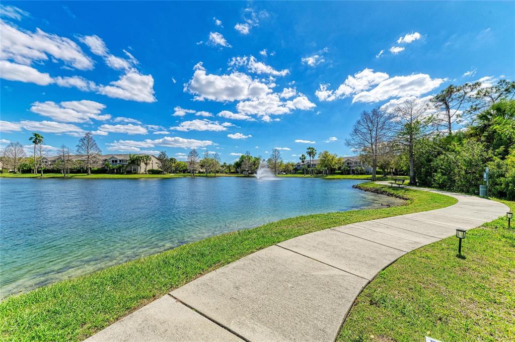 A concrete path curves along a lake with a fountain and apartment buildings in background — 8911 MANOR LOOP UNIT 207, LAKEWOOD RANCH, FL