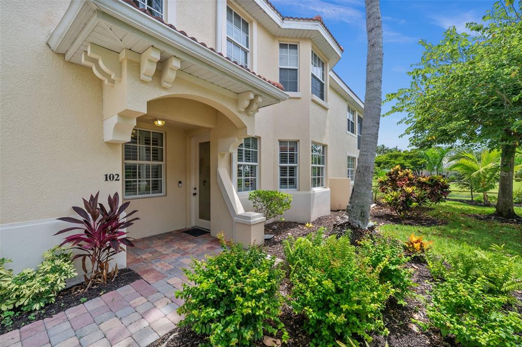 The exterior of a two-story tan stucco building features a covered entryway and brick walkway — 5626 CAPE HARBOUR DRIVE UNIT 102, CAPE CORAL, FL