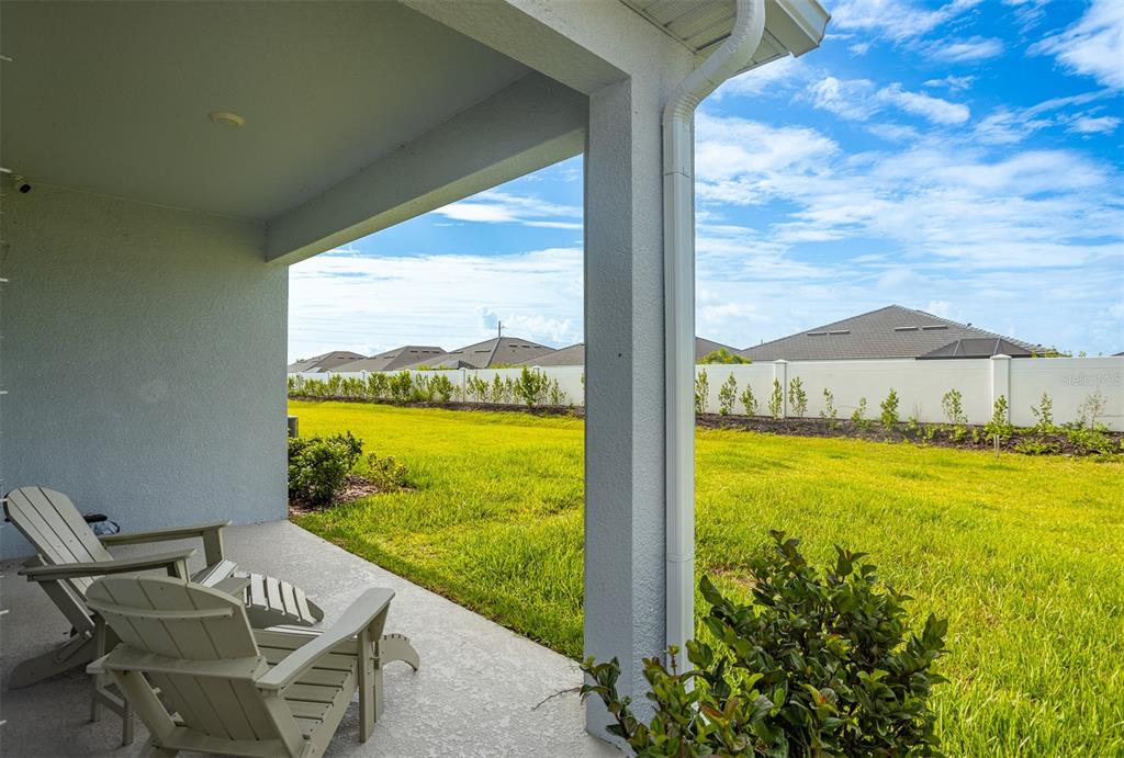 A covered concrete patio features two Adirondack chairs overlooking a grassy backyard with a fence — 18052 CROPSIDE TRAIL, BRADENTON, FL