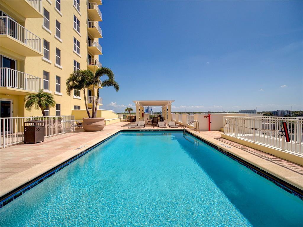 A rectangular swimming pool sits on a terrace next to a tall yellow apartment building — 628 CLEVELAND STREET UNIT 1314, CLEARWATER, FL