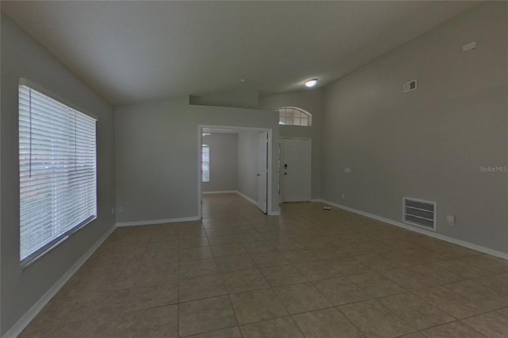 An empty living room features light-colored tile flooring, vaulted ceilings, and a white front door — 13809 RIVERPATH GROVE DRIVE, ORLANDO, FL