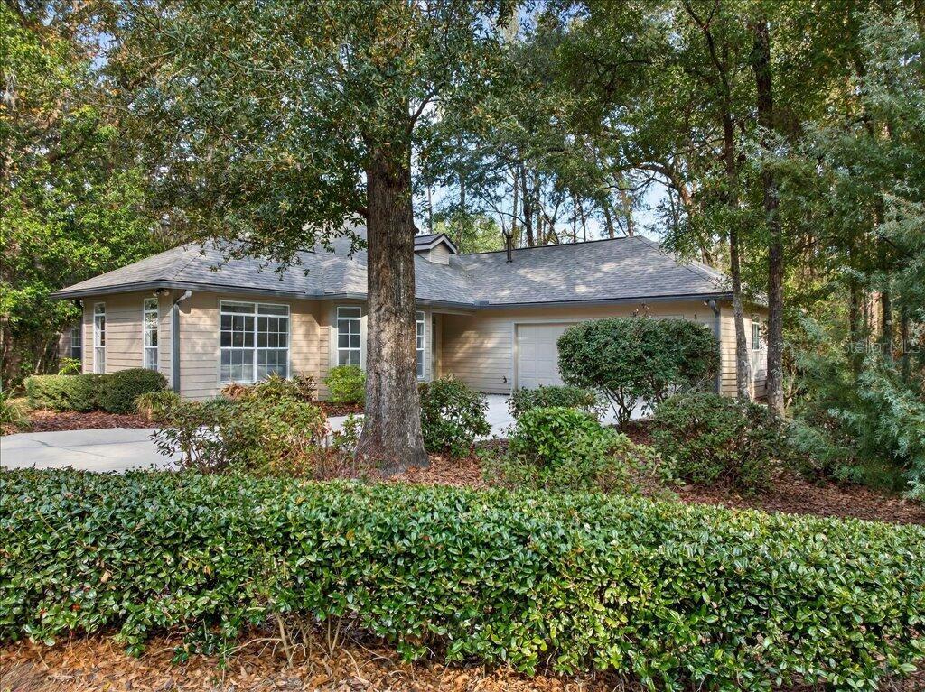 A tan single-story house with a two-car garage sits behind a manicured hedge and trees — 4412 SW 101ST DRIVE, GAINESVILLE, FL