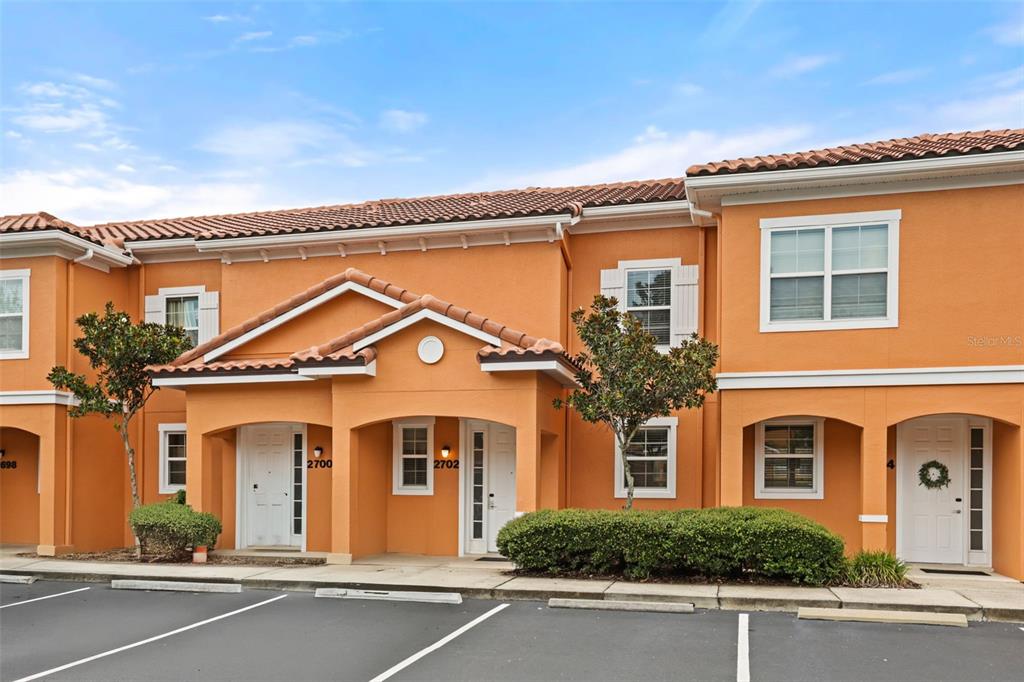 An exterior view of a two-story orange stucco townhouse building with three white front doors — 2702 CORVETTE LANE, KISSIMMEE, FL