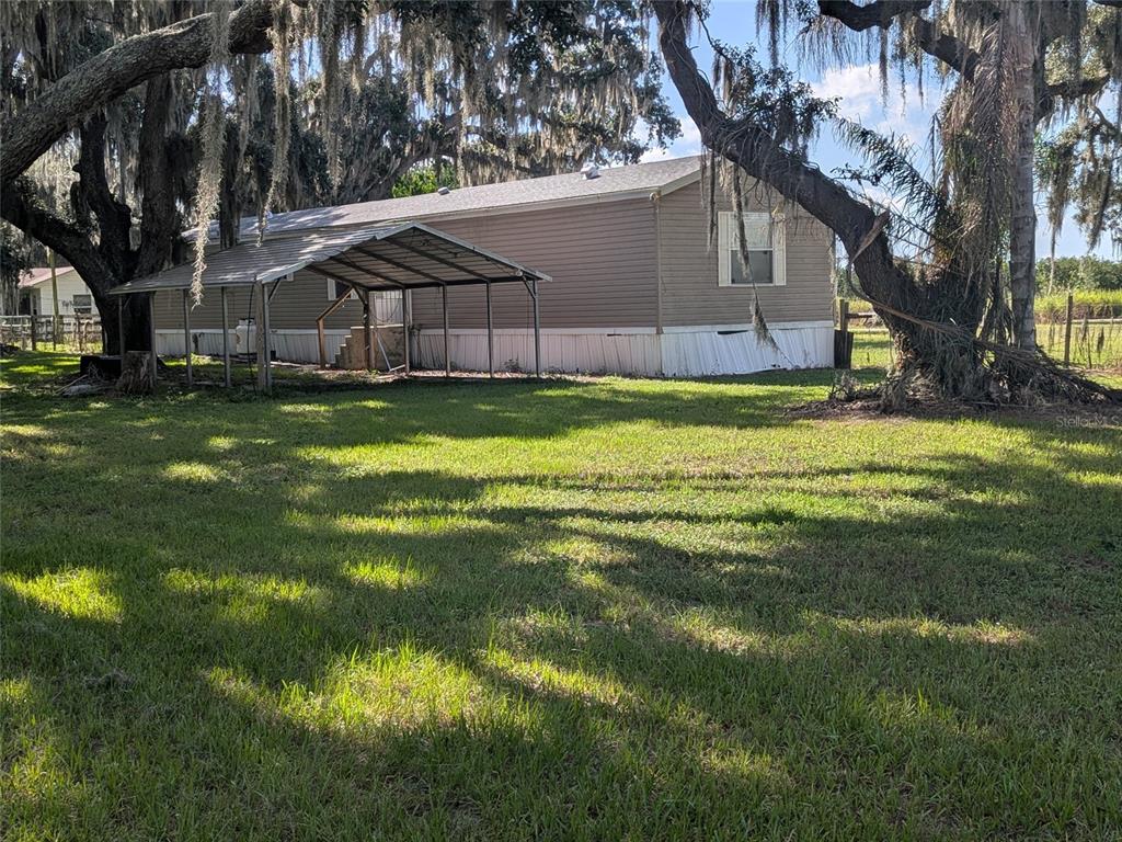 A single-story manufactured home with a metal carport sits on a grassy lot under trees — 3193 NE DAVIS STREET, ARCADIA, FL