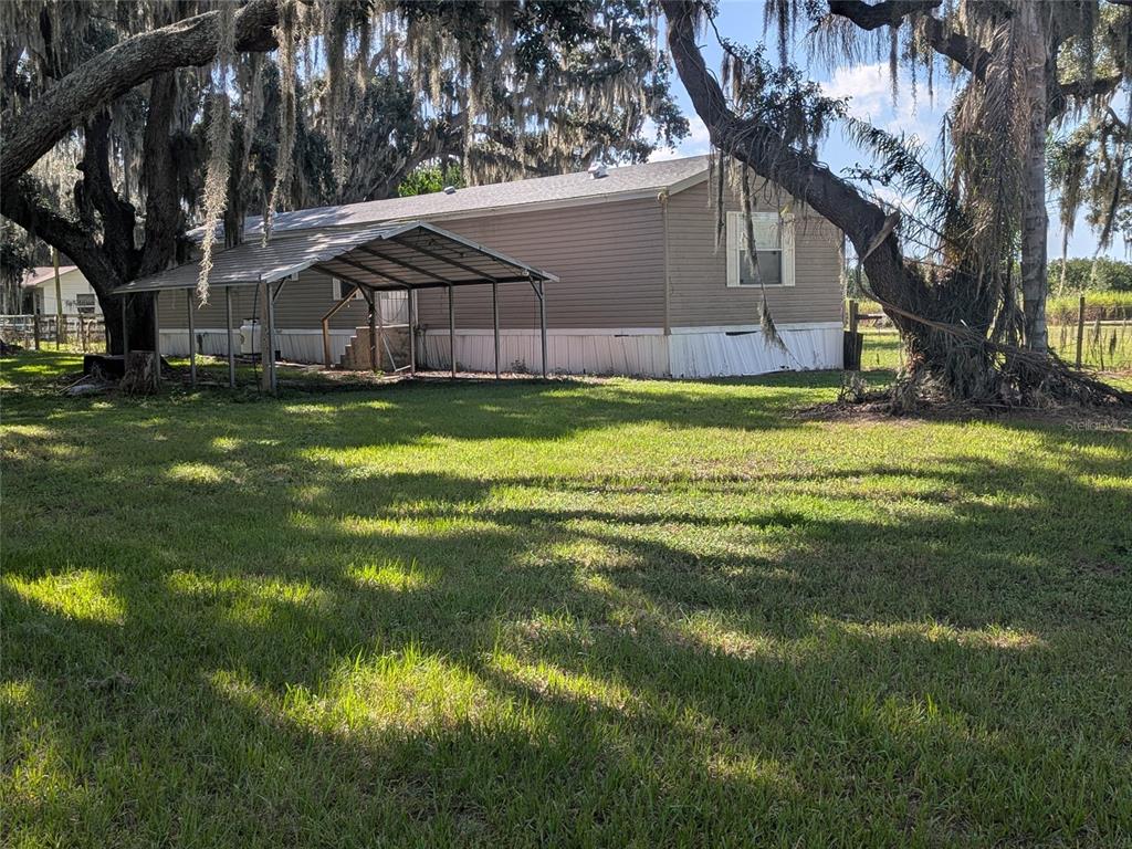 A tan manufactured home with a metal carport sits on a grassy lot under trees — 3193 NE DAVIS STREET, ARCADIA, FL