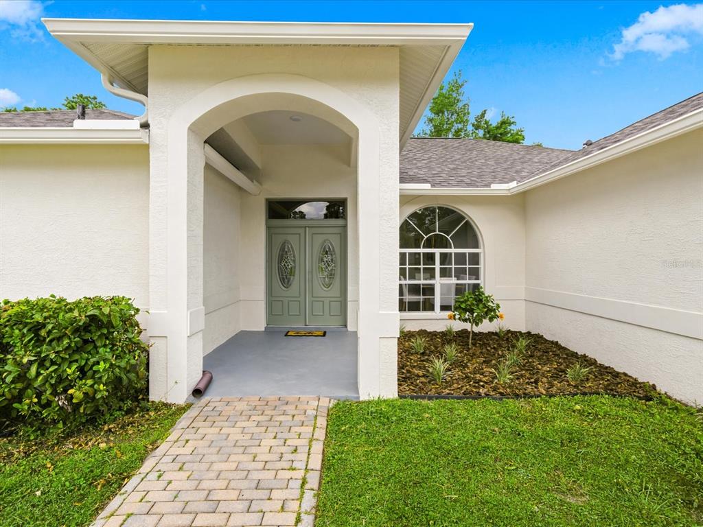 A white stucco house exterior features a covered arched entryway with sage green double doors — 4617 DEBBIE LANE, LUTZ, FL